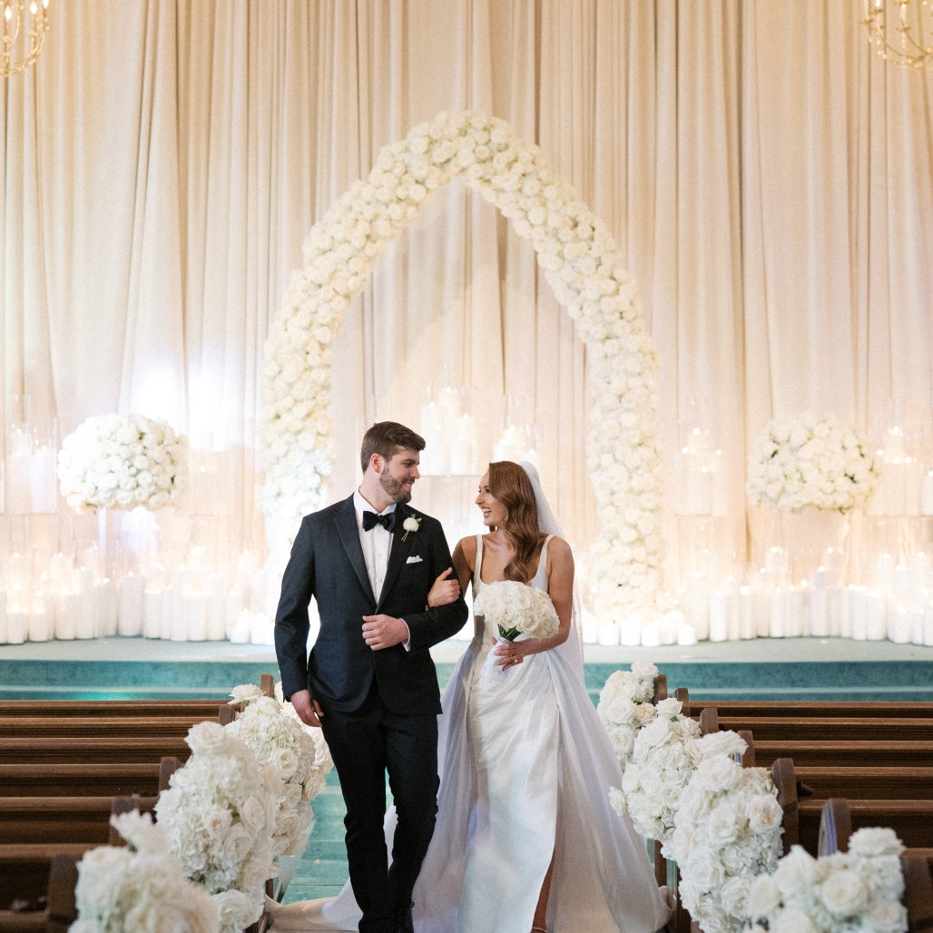 Bride and groom beneath a white floral arch lined with pillar candles
